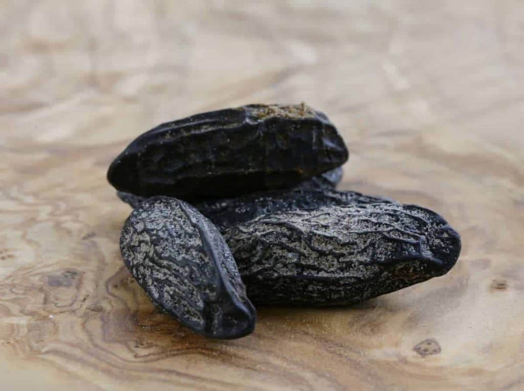 Drei getrocknete Tonkabohnen liegen auf einer glatten, hellen Holzoberfläche mit sichtbarer Maserung. Die Bohnen sind dunkel, runzelig und haben eine leicht glänzende Oberfläche.