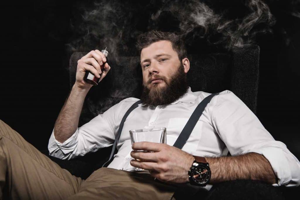 Ein bärtiger Mann in einem weißen Hemd und Hosenträgern sitzt auf einem Stuhl, hält in der einen Hand ein E-Zigaretten-Gerät und in der anderen ein Glas Whisky, während vor einem dunklen Hintergrund Rauch um ihn herumwirbelt.