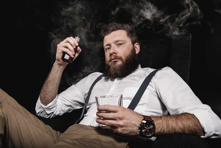 Ein bärtiger Mann in einem weißen Hemd und Hosenträgern sitzt auf einem Stuhl, hält in der einen Hand ein E-Zigaretten-Gerät und in der anderen ein Glas Whisky, während vor einem dunklen Hintergrund Rauch um ihn herumwirbelt.
