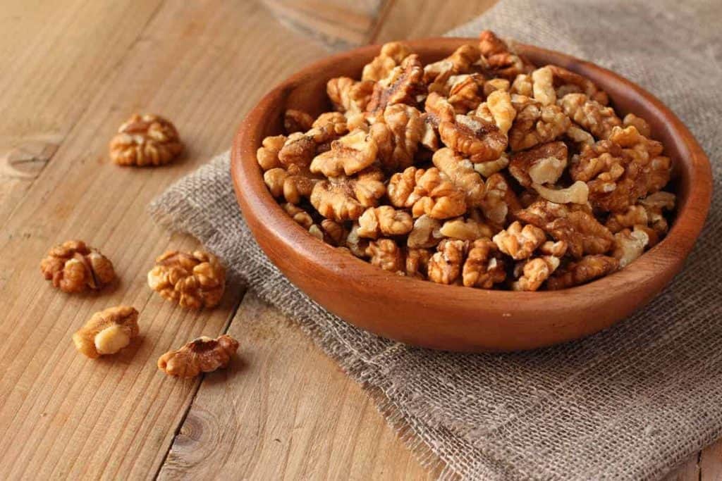 Auf einem Stück Sackleinen steht eine Holzschale, gefüllt mit geschälten Walnusshälften, und auf einem Holztisch daneben liegen ein paar Walnüsse verstreut.