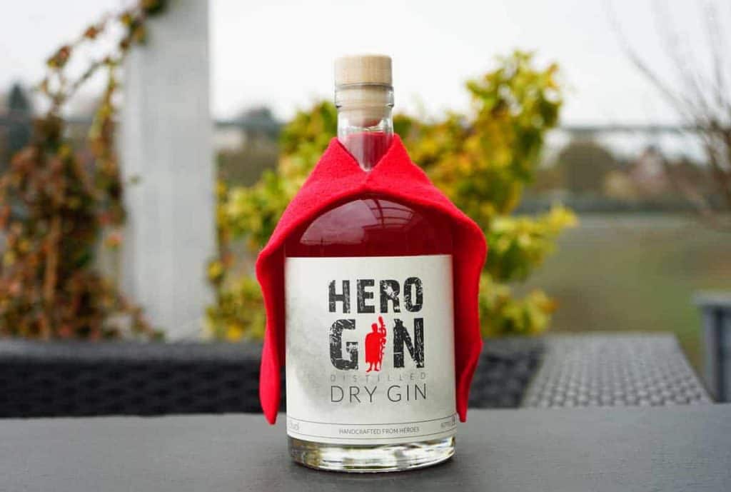 Eine Flasche Hero Gin, über deren Schultern ein roter Umhang drapiert ist, steht im Freien auf einem dunklen Tisch, im Hintergrund ist verschwommenes Grün zu sehen.