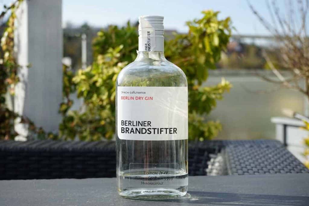 Auf einem Tisch im Freien steht eine durchsichtige Flasche „Berliner Brandstifter Berlin Dry Gin“, umgeben von grünem Laub und vor dem verschwommenen Hintergrund eines Geländers und von Gebäuden.