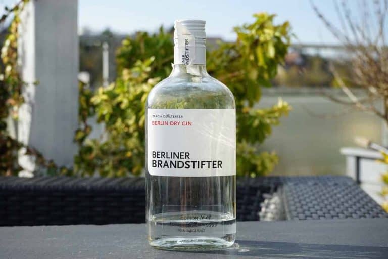 Auf einem Tisch im Freien steht eine durchsichtige Flasche „Berliner Brandstifter Berlin Dry Gin“, umgeben von grünem Laub und vor dem verschwommenen Hintergrund eines Geländers und von Gebäuden.