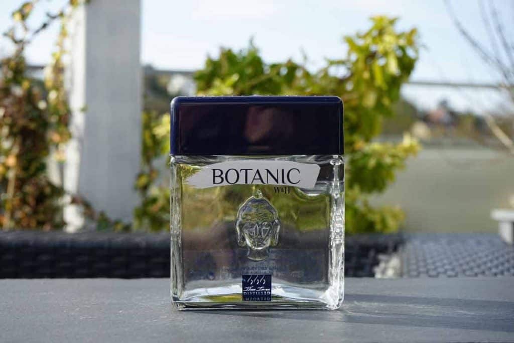 Auf einem Tisch im Freien steht eine durchsichtige Glasflasche mit der Aufschrift „BOTANIC“ und einem dunkelblauen Verschluss; dahinter sind grünes Laub und ein unscharfer Hintergrund zu sehen.
