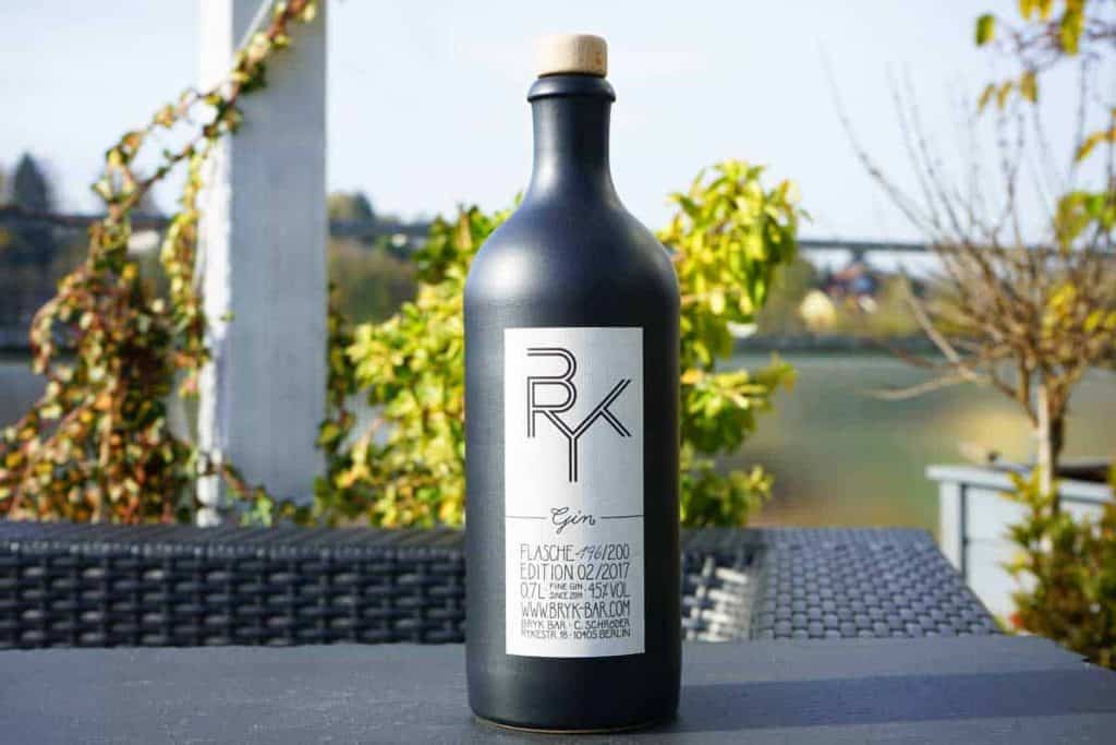 Eine mattschwarze Gin-Flasche mit dem Etikett „BRYX Gin“ steht auf einem Terrassentisch im Freien, umgeben von grünen Pflanzen und einem verschwommenen Naturhintergrund.