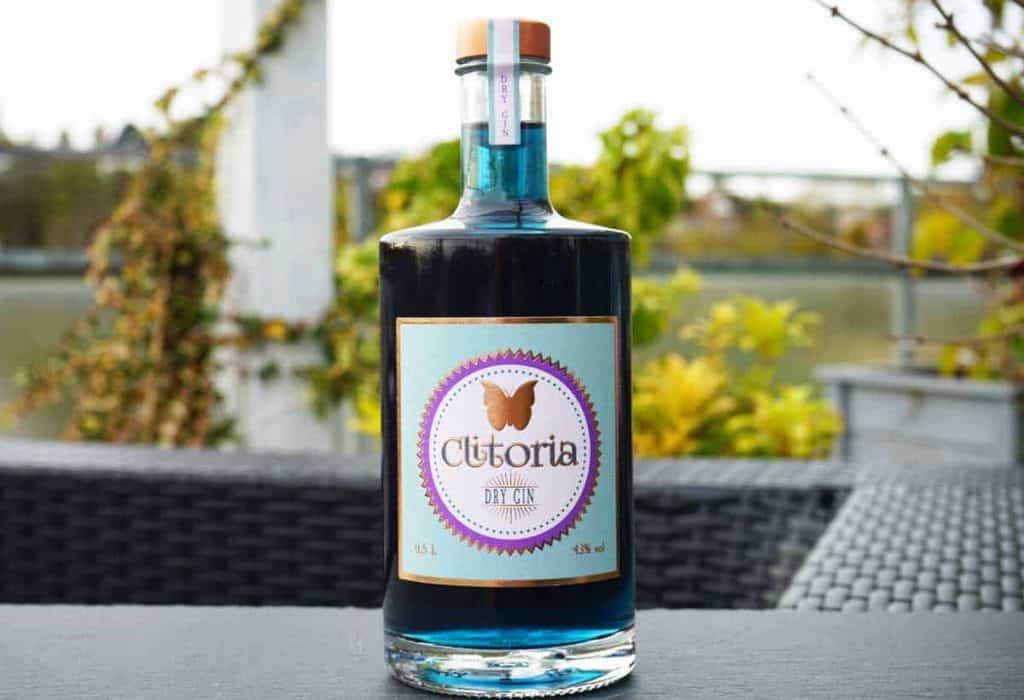 Auf einem Tisch im Freien steht eine Flasche Clitoria Dry Gin mit blauer Flüssigkeit darin, im Hintergrund sind Pflanzen und Wasser verschwommen zu sehen.