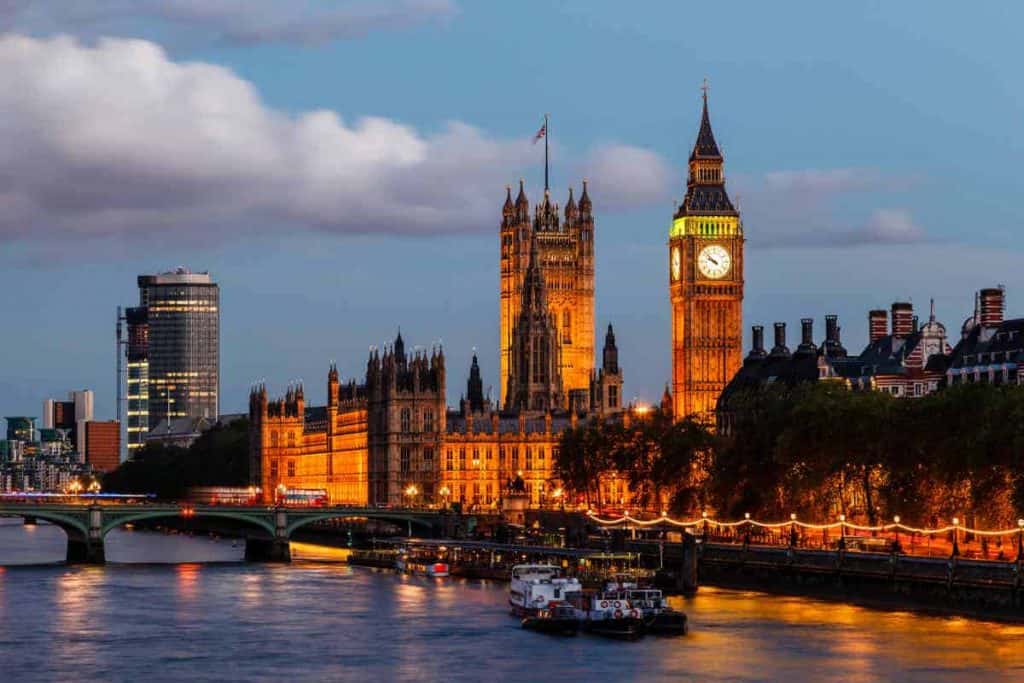 Der beleuchtete Big Ben und die Houses of Parliament an der Themse in London bei Einbruch der Dämmerung, mit der Westminster Bridge und Booten im Vordergrund.