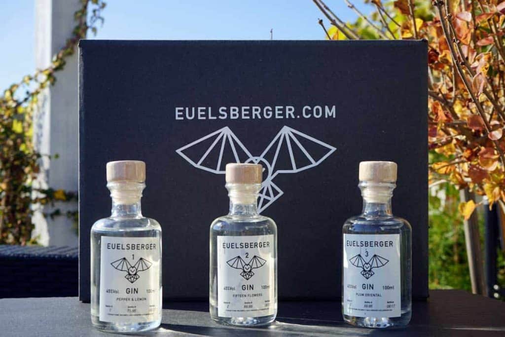 Drei kleine Flaschen Euelsberger Gin stehen vor einer schwarzen Schachtel, auf der ein geometrisches Fledermaus-Logo und der Schriftzug EUELSBERGER.COM zu sehen sind. Im Hintergrund sind herbstliche Zweige zu erkennen.