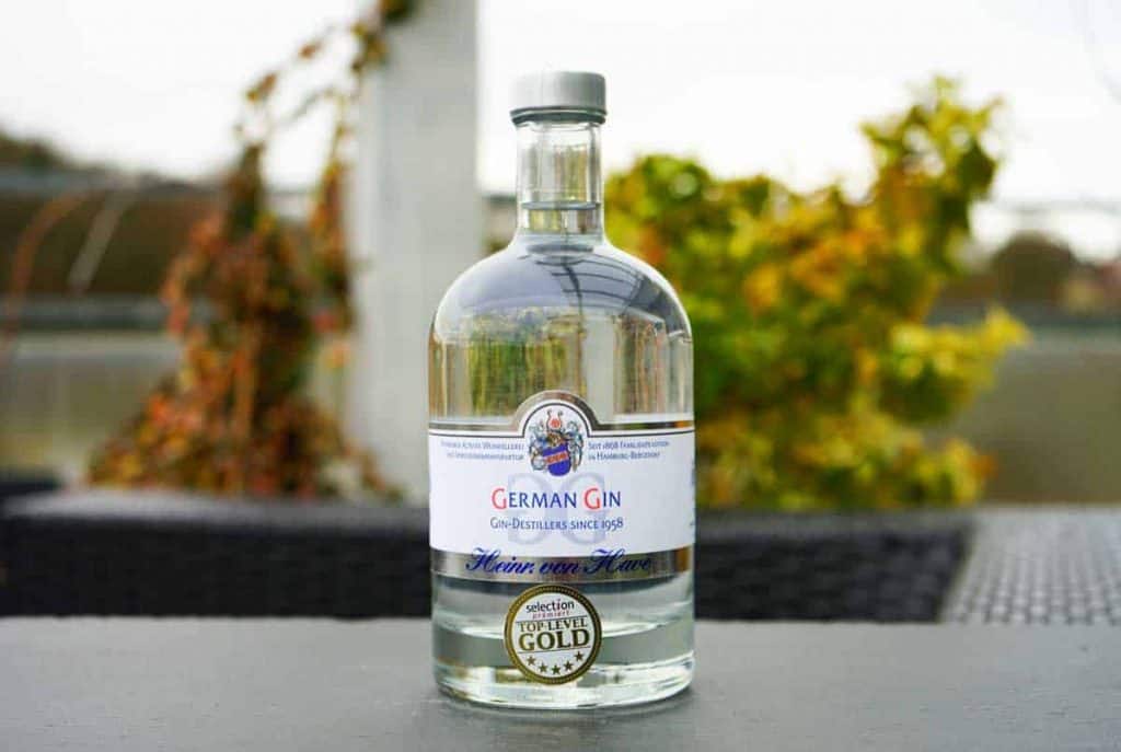 Auf einem schwarzen Tisch im Freien steht eine durchsichtige Flasche mit deutschem Gin und einem weißen Etikett, im Hintergrund ist verschwommenes grünes und gelbes Laub zu sehen.