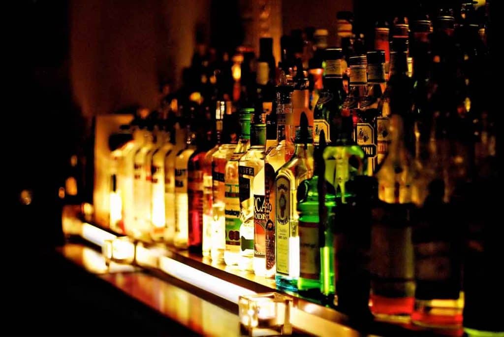 Auf einem schwach beleuchteten Barregal stehen Flaschen mit verschiedenen alkoholischen Getränken in Reih und Glied, beleuchtet von warmem, stimmungsvollem Licht, vor denen einige kleine Kerzen stehen.