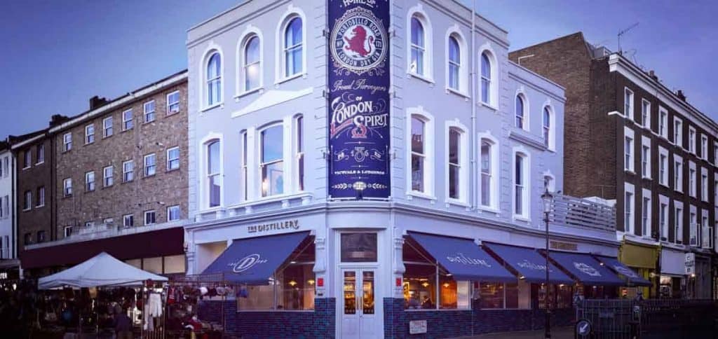 In einem weißen Eckgebäude mit großen Fenstern und blauen Markisen befindet sich „The Distillery“ an einer belebten Straße mit Marktständen und Menschen. Auf einem hohen Schild steht „The London Spirit“. Das Abendlicht taucht die Szene in einen violett-blauen Farbton.