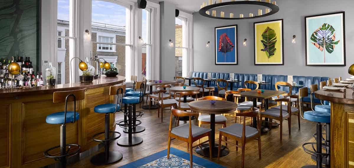 Moderne Bar mit Holztischen, blauen gepolsterten Stühlen, einer geschwungenen Holztheke und großen Fenstern. An der grauen Wand über einer langen blauen Bank hängen drei farbenfrohe Blattmotive.