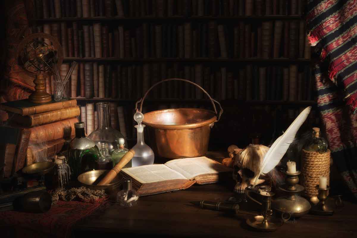 Auf einem schwach beleuchteten Schreibtisch liegen alte Bücher, Glasflaschen, eine Feder, ein Schädel, ein Kupfereimer und eine Kerze; im Hintergrund sind Bücherregale und ein gemustertes Tuch zu sehen.