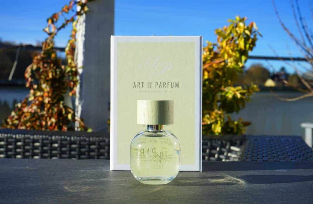 Auf einem Tisch im Freien, im Hintergrund sind verschwommene Pflanzen und ein blauer Himmel zu sehen, steht ein durchsichtiger Parfümflakon mit goldenem Verschluss vor einer passenden Schachtel mit der Aufschrift „Art de Parfum“.
