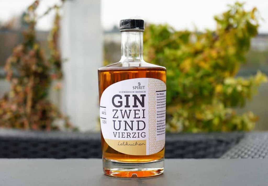 Eine durchsichtige Flasche „Gin Zweiundvierzig Lebkuchen“ steht im Freien auf einer dunklen Fläche, im Hintergrund ist grünes Laub verschwommen zu erkennen.