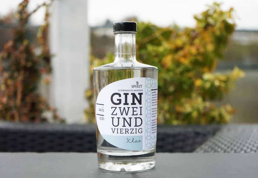 Auf einem dunklen Tisch im Freien steht eine durchsichtige Gin-Flasche mit der Aufschrift „Gin Zweiundvierzig“, im Hintergrund sind verschwommene Pflanzen und ein weißer Pfosten zu sehen.