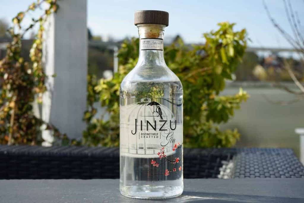 Auf einem dunklen Tisch im Freien steht eine durchsichtige Jinzu-Gin-Flasche mit einem Holzverschluss, im Hintergrund sind grüne Pflanzen und verschwommene Gebäude zu sehen.