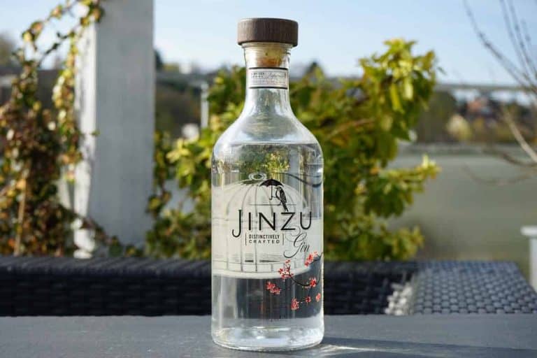 Auf einem dunklen Tisch im Freien steht eine durchsichtige Jinzu-Gin-Flasche mit einem Holzverschluss, im Hintergrund sind grüne Pflanzen und verschwommene Gebäude zu sehen.