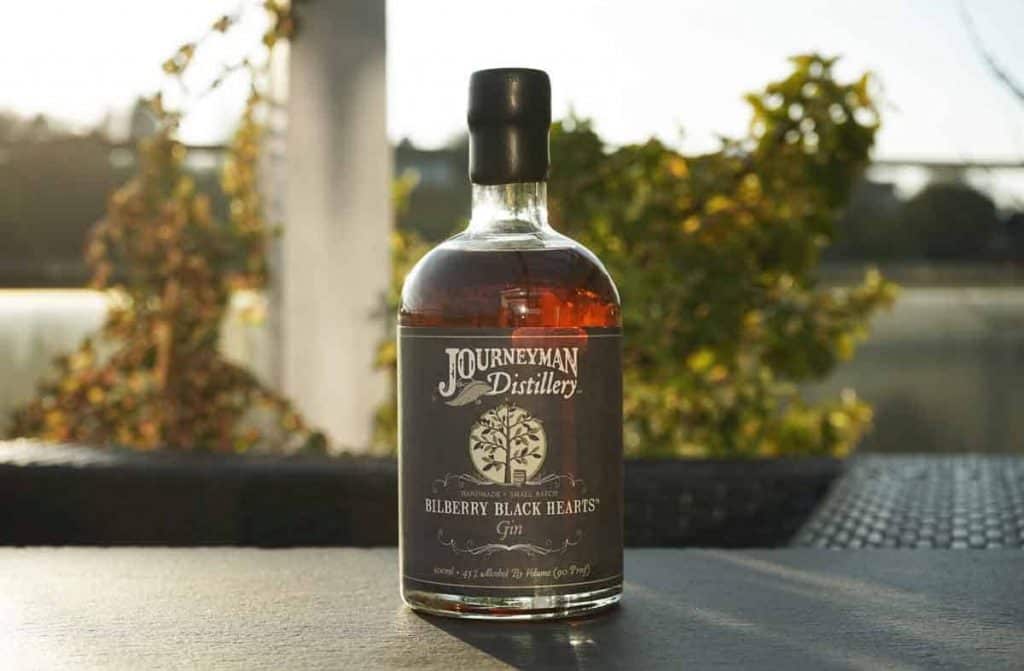 Auf einem dunklen Tisch im Freien steht eine Flasche „Blueberry Black Hearts Gin“ von der Journeyman Distillery, im Hintergrund sind verschwommene Grünpflanzen und Sonnenlicht zu sehen.