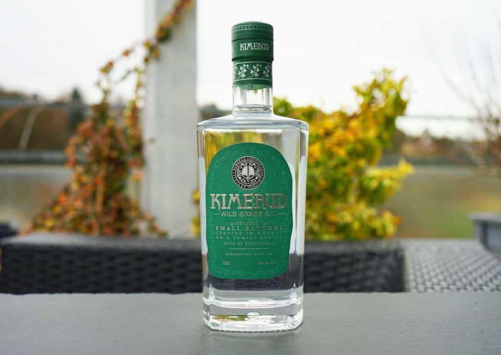 Eine Flasche „Kimerud Wild Grade Gin“ mit grünem Etikett steht auf einem dunklen Tisch im Freien, im Hintergrund sind verschwommene Herbstblätter und ein Gewässer zu sehen.