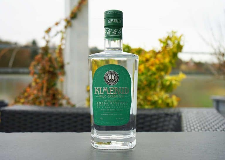 Eine Flasche „Kimerud Wild Grade Gin“ mit grünem Etikett steht auf einem dunklen Tisch im Freien, im Hintergrund sind verschwommene Herbstblätter und ein Gewässer zu sehen.