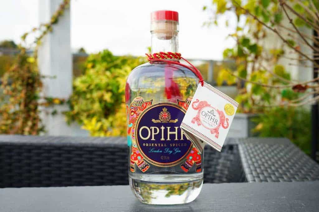 Auf einem schwarzen Gartentisch steht eine Flasche „Opihr Oriental Spiced London Dry Gin“, umgeben von Grünpflanzen und vor einem unscharfen Hintergrund. Am Flaschenhals ist ein rot-goldenes Etikett befestigt.
