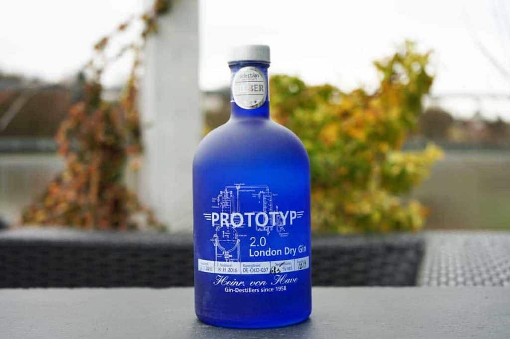 Auf einem schwarzen Tisch im Freien steht eine blaue Flasche PROTOTYP 2.0 London Dry Gin, im Hintergrund sind verschwommene Pflanzen und ein weißer Pfosten zu sehen.
