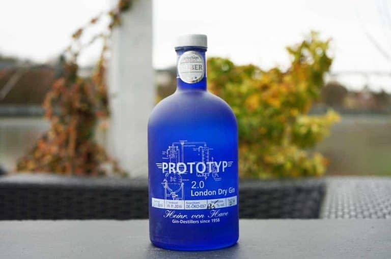 Auf einem schwarzen Tisch im Freien steht eine blaue Flasche PROTOTYP 2.0 London Dry Gin, im Hintergrund sind verschwommene Pflanzen und ein weißer Pfosten zu sehen.