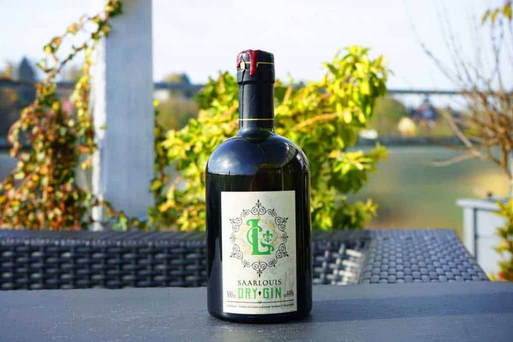 Auf einem schwarzen Gartentisch steht an einem sonnigen Tag eine Flasche Saarlouis Dry Gin, im Hintergrund sind Grünpflanzen und eine verschwommene Landschaft zu sehen.
