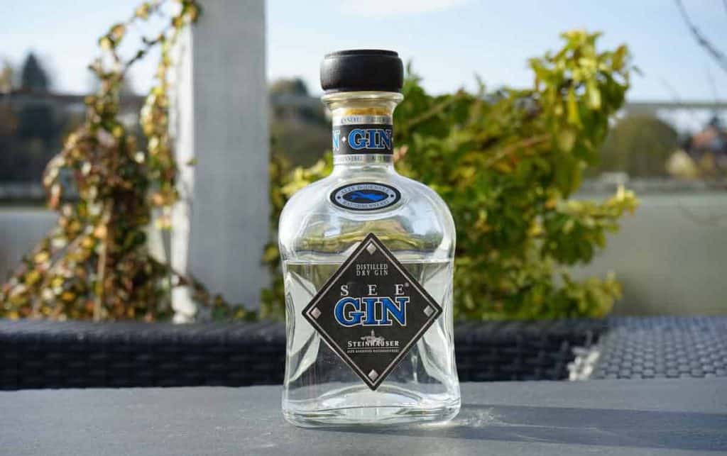Auf einem Tisch im Freien steht eine durchsichtige Glasflasche „See Gin“ mit schwarzem Verschluss. Dahinter sind an einem sonnigen Tag grünes Laub und ein verschwommener Hintergrund zu sehen.