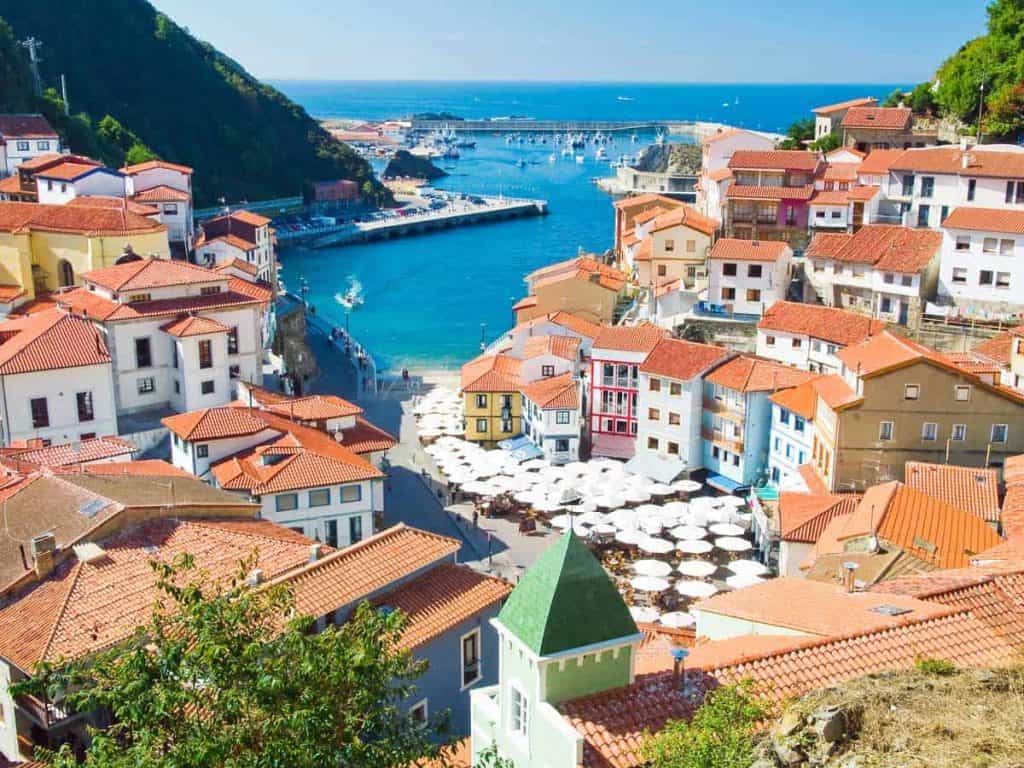 Ein farbenfrohes Küstendorf mit roten Ziegeldächern und Blick auf einen blauen Hafen, Boote, die im Yachthafen vor Anker liegen, üppig grüne Hügel und weiße Sonnenschirme, die unter klarem Himmel einen Terrassenbereich nahe der Uferpromenade überdachen.