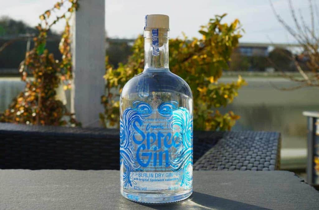 Auf einem Tisch im Freien steht eine Flasche Spre Gin, im Hintergrund sind Grünpflanzen und ein Gewässer zu sehen. Das Sonnenlicht spiegelt sich in der durchsichtigen Flasche und bringt das blaue Etikett und das aufwendige Design besonders gut zur Geltung.