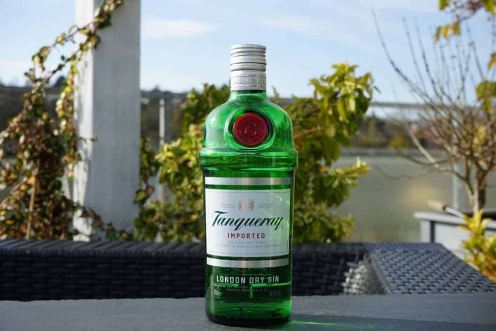 Auf einem schwarzen Gartentisch steht eine grüne Flasche Tanqueray London Dry Gin, im Hintergrund sind Pflanzen und Wasser verschwommen zu sehen.