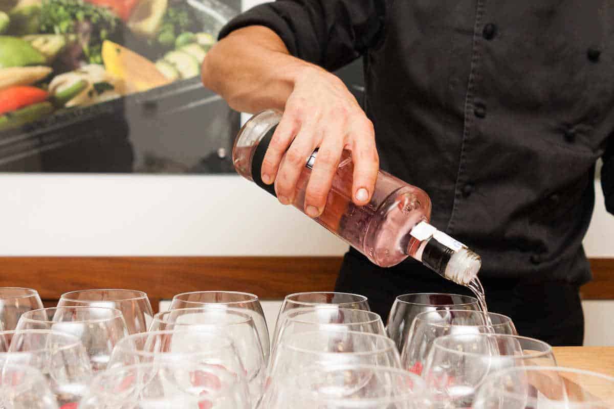 Eine Person in einem schwarzen Hemd schenkt Roséwein aus einer Flasche in mehrere leere Weingläser ein, die auf einem Tisch stehen.