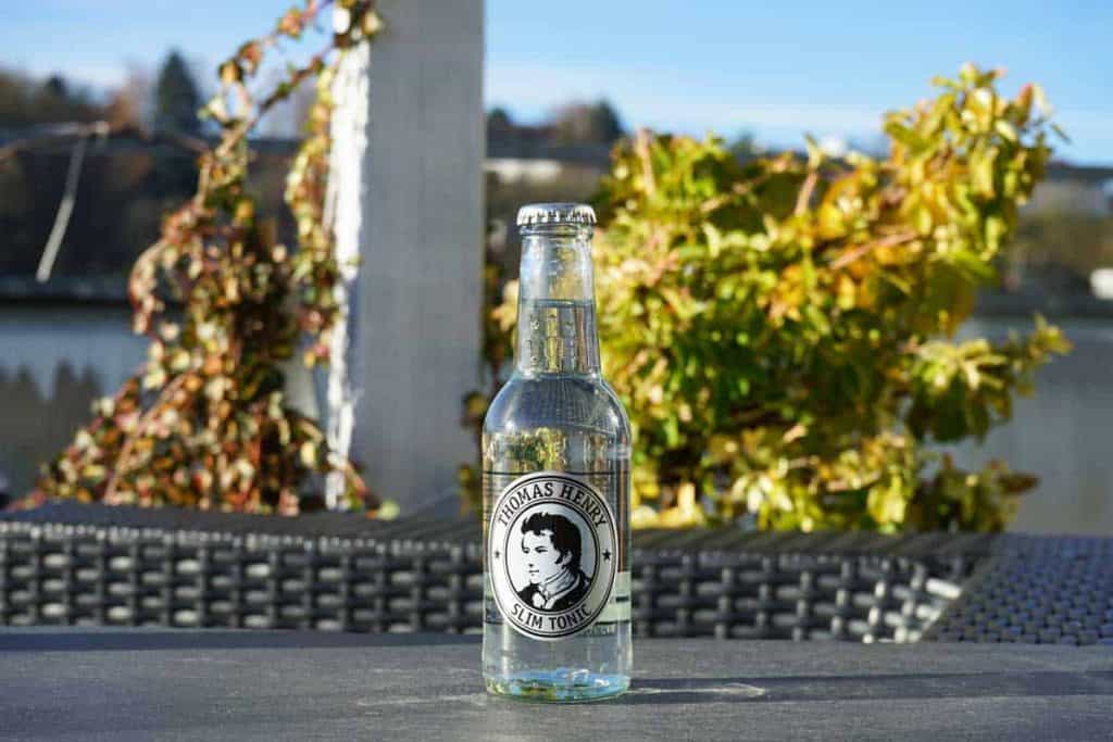 Auf einem Tisch im Freien steht eine klare Glasflasche mit „Thomas Henry Slim Tonic“; im hellen Tageslicht sind grüne Pflanzen und ein verschwommener Hintergrund zu sehen.