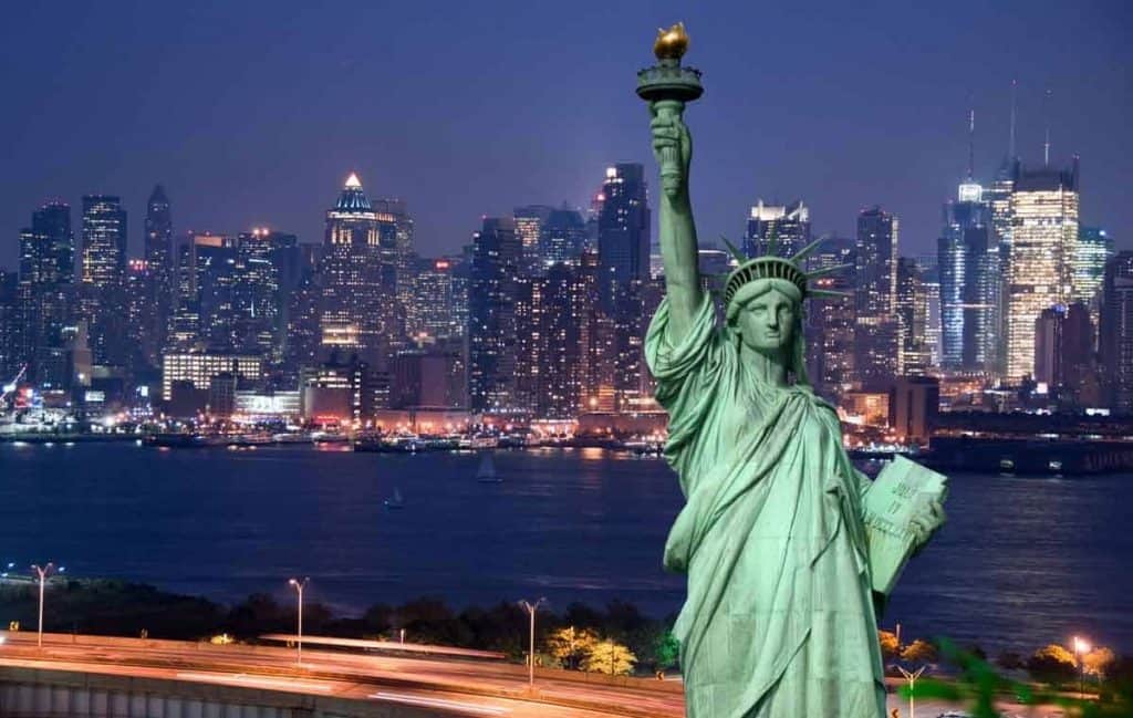 Im Vordergrund steht die Freiheitsstatue, und auf der anderen Seite des Wassers ist nachts die beleuchtete Skyline von New York City zu sehen.