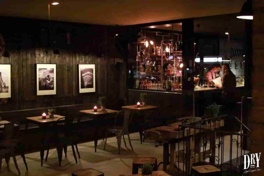Eine schwach beleuchtete Bar mit kleinen Tischen und Stühlen, Kerzen auf jedem Tisch, gerahmten Fotos an einer dunklen Wand und einem gut sortierten Barbereich mit Flaschen und Gläsern. Hinter der Bar ist eine Person bei der Arbeit. DRY-Logo in der Ecke.