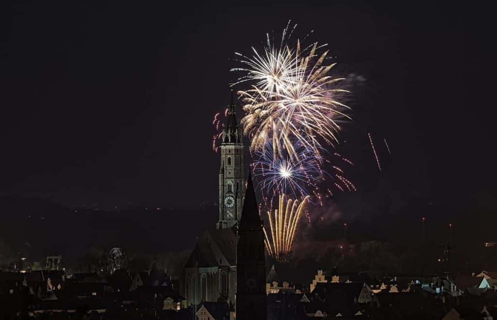 Hinter einem großen Uhrturm und einer Kirche explodieren Feuerwerkskörper am Nachthimmel und beleuchten die Dächer der darunter liegenden Stadt.