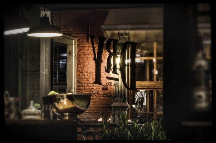 Ein warm beleuchtetes Bar-Interieur mit Backsteinwänden, einer großen, spiegelnden Schale auf der Theke, hängenden Lampen und dem Schriftzug „DRY“, der spiegelverkehrt auf die Fensterscheibe geschrieben ist.