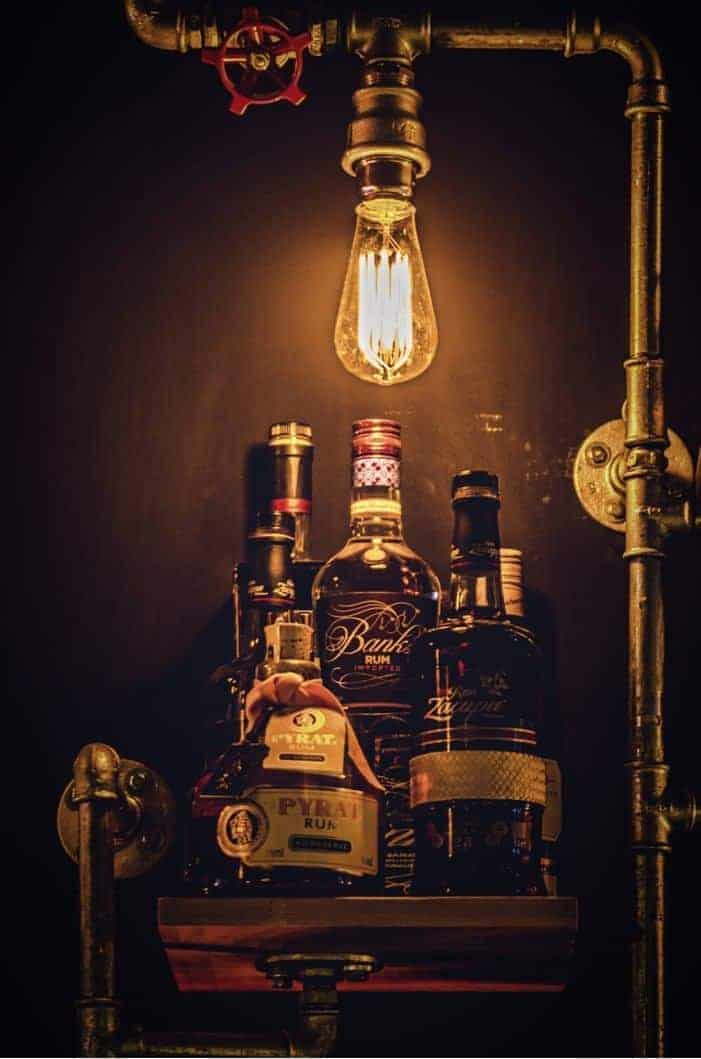 Ein schwach beleuchtetes Regal mit Alkoholflaschen unter einer alten, freiliegenden Glühbirne, umgeben von Metallrohren im Industrial-Stil vor einem dunklen Hintergrund.
