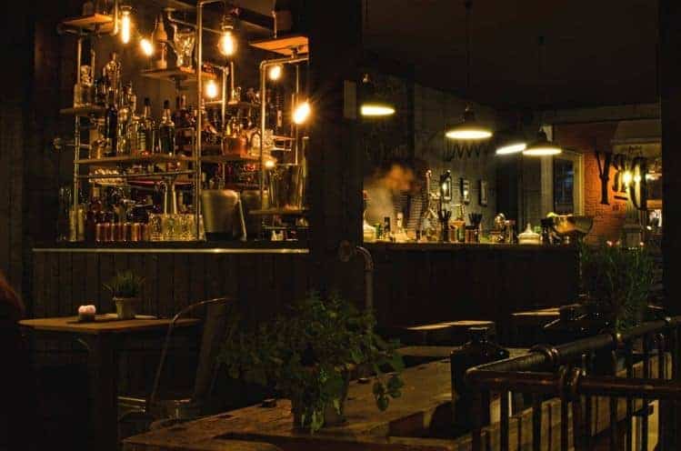 Eine schummrig beleuchtete Bar mit Hängelampen, Flaschen in den Regalen und einem verschwommenen Barkeeper hinter der Theke. In dem gemütlichen, rustikalen Ambiente sind Holztische und -stühle zu sehen.