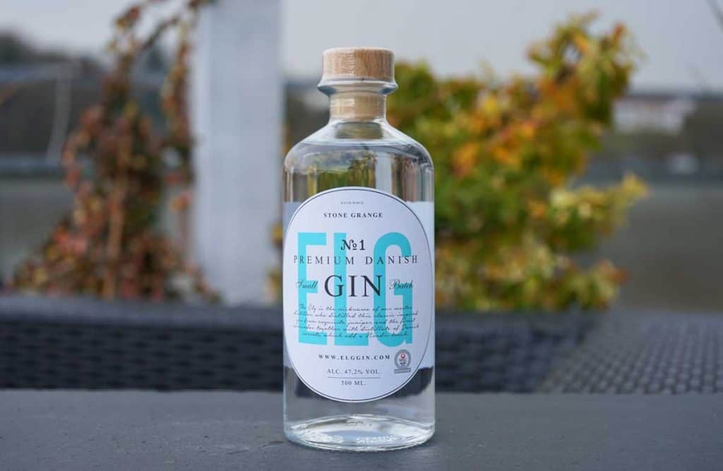 Eine durchsichtige Flasche „Stone Grange No. 1 Premium Danish Gin“ mit einem Holzverschluss steht im Freien auf einer dunklen Oberfläche, im Hintergrund sind verschwommene Grünpflanzen und ein Gewässer zu sehen.
