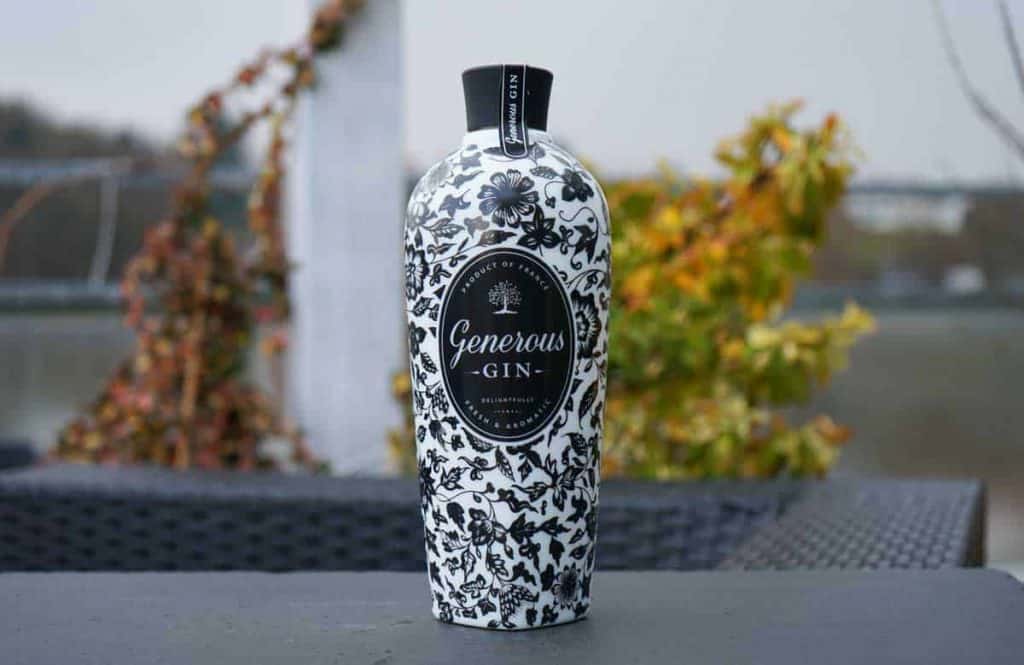 Auf einem grauen Tisch im Freien steht eine weiße Flasche „Generous Gin“ mit schwarzen Blumenmustern, im Hintergrund sind verschwommene Pflanzen und ein Geländer zu sehen.