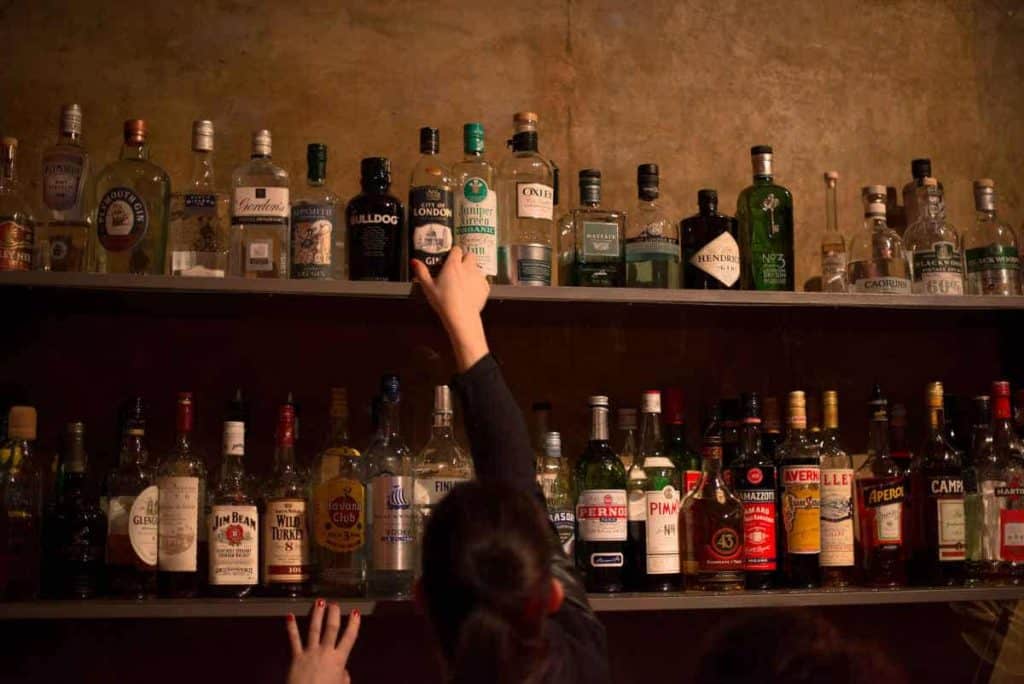 Eine Person greift nach einer Flasche Schnaps im obersten Regal einer schummrig beleuchteten Bar, umgeben von verschiedenen Alkoholflaschen, die auf zwei Regalen aufgereiht sind.