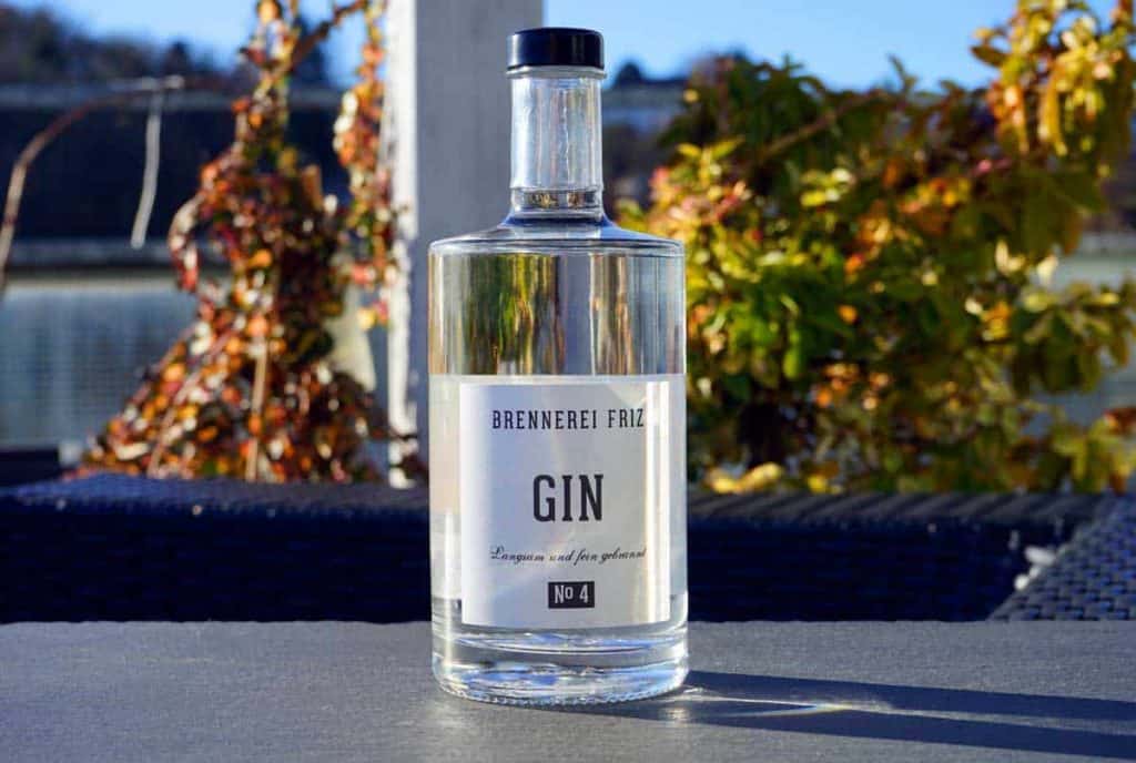 Auf einem Tisch im Freien steht eine Gin-Flasche aus klarem Glas mit der Aufschrift „Brennerei Fritz Gin No 4“, im Hintergrund sind verschwommene Herbstblätter und Sonnenlicht zu sehen.