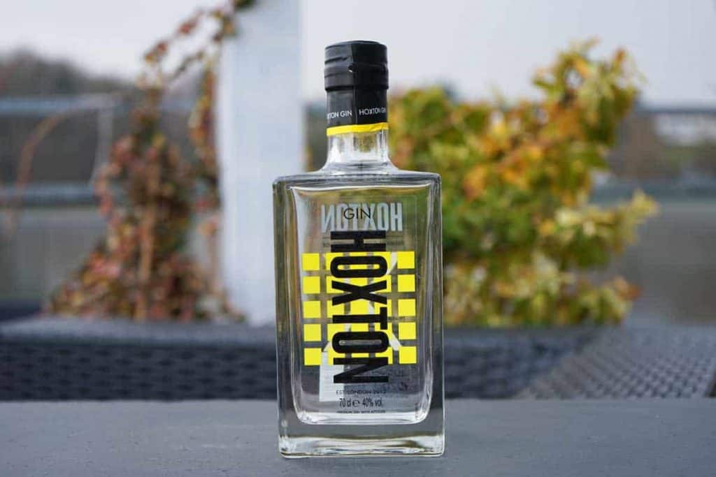 Eine klare Glasflasche mit Hoxton-Gin steht im Freien auf einer grauen Fläche, im Hintergrund sind Pflanzen und verschwommenes Grün zu sehen. Das Etikett ist mit auffälligen gelben und schwarzen geometrischen Mustern verziert.