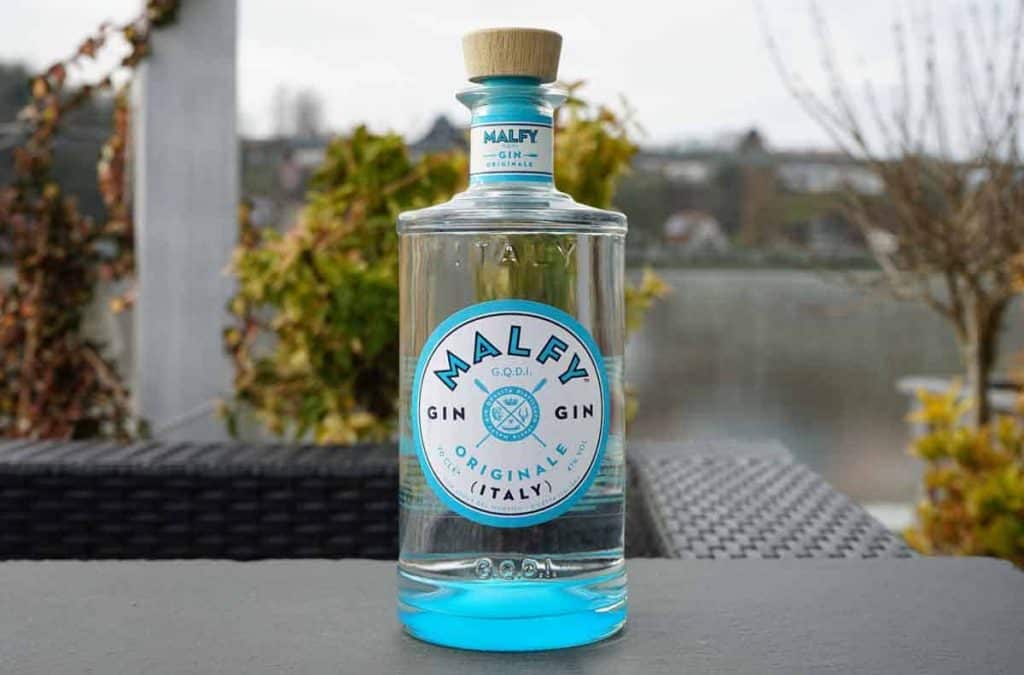 Auf einem dunklen Tisch im Freien steht eine durchsichtige Glasflasche Malfy Gin mit einem blau-weißen Etikett, im Hintergrund sind Grünpflanzen und verschwommene Gebäude zu sehen.