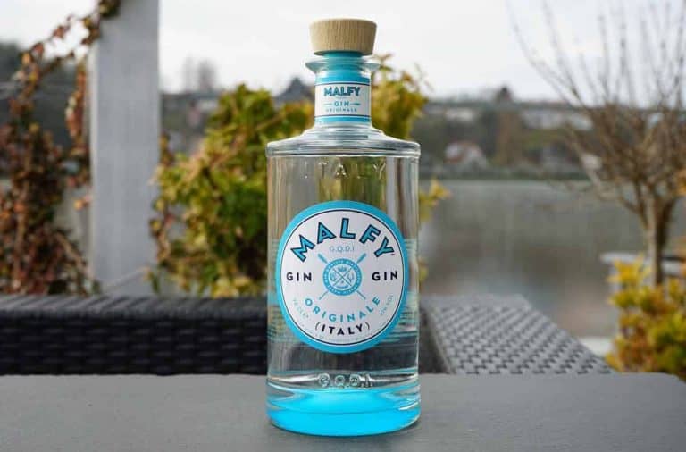 Auf einem dunklen Tisch im Freien steht eine durchsichtige Glasflasche Malfy Gin mit einem blau-weißen Etikett, im Hintergrund sind Grünpflanzen und verschwommene Gebäude zu sehen.