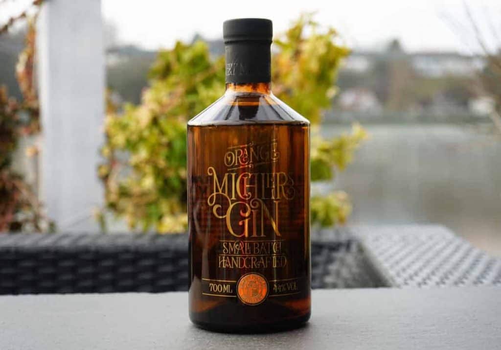Auf einem Tisch im Freien steht eine braune Flasche Michlers Gin, dahinter sind grünes Laub und ein unscharfer Hintergrund zu sehen.