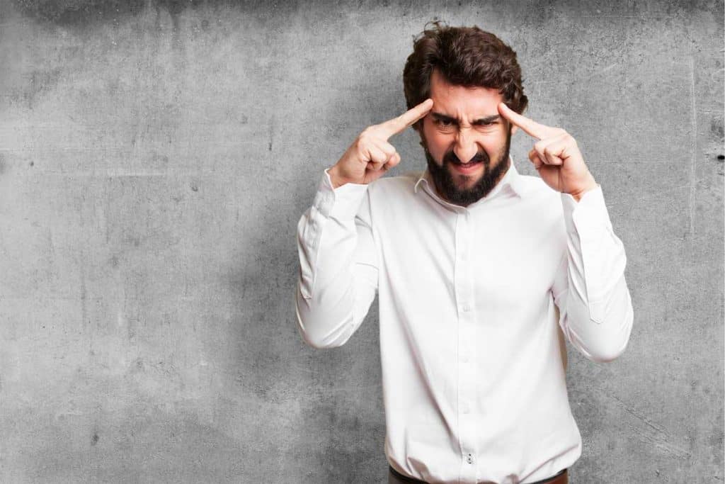 Ein Mann in einem weißen Hemd steht vor einer grauen Betonwand, legt beide Zeigefinger an die Schläfen und kneift die Augen zusammen; er wirkt tief in Gedanken versunken oder hochkonzentriert.